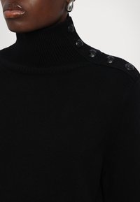 Zwarte gebreide coltrui met ribbelstructuur en vijf decoratieve zwarte knopen op de schouder voor extra detail.