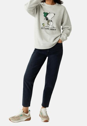 Vrouw draagt een lichtgrijze trui met Snoopy- en Woodstock-graphic en de tekst "Out and About", donkere spijkerbroek en beige sneakers met turquoise accenten.