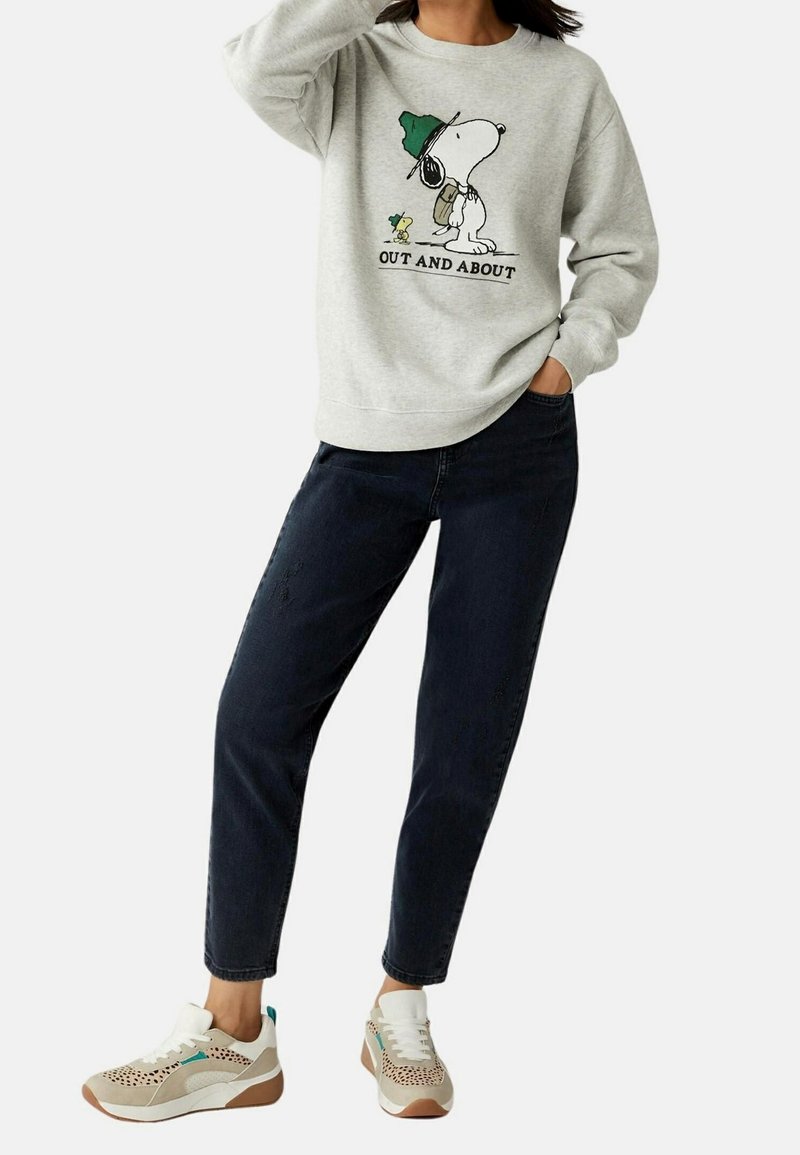 Femme portant un sweat-shirt gris clair avec un graphisme Snoopy et Woodstock et le texte « Out and About », jean foncé et baskets beiges avec des accents turquoise.