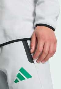 Jasnoszary strój sportowy Adidas z czarnymi akcentami i zielonym logo. Zbliżenie na dłoń spoczywającą na kieszeni z suwakiem. Gładka tekstura.