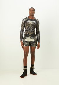 Long-sleeve graphic athletic top with black and gold designs; paired with black shorts featuring a bold pattern. Worn with black athletic shoes.