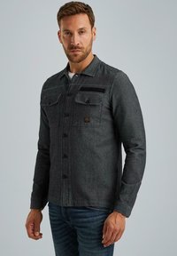 Camicia in denim grigio con colletto, chiusura con bottoni e due tasche sul petto. Presenta un tessuto testurizzato e una piccola etichetta del marchio.