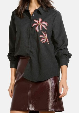 Femme portant une chemise gris foncé à boutons ornée de fleurs roses brodées et une jupe en cuir brun foncé brillant, debout les mains détendues.