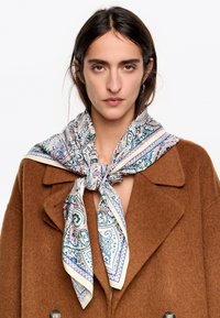 Jedwabny szalik w wzór paisley w kolorach niebieskim, różowym i kremowym, przewiązany wokół szyi na brązowym wełnianym płaszczu. Z wykończeniem w kremowej bordurze.