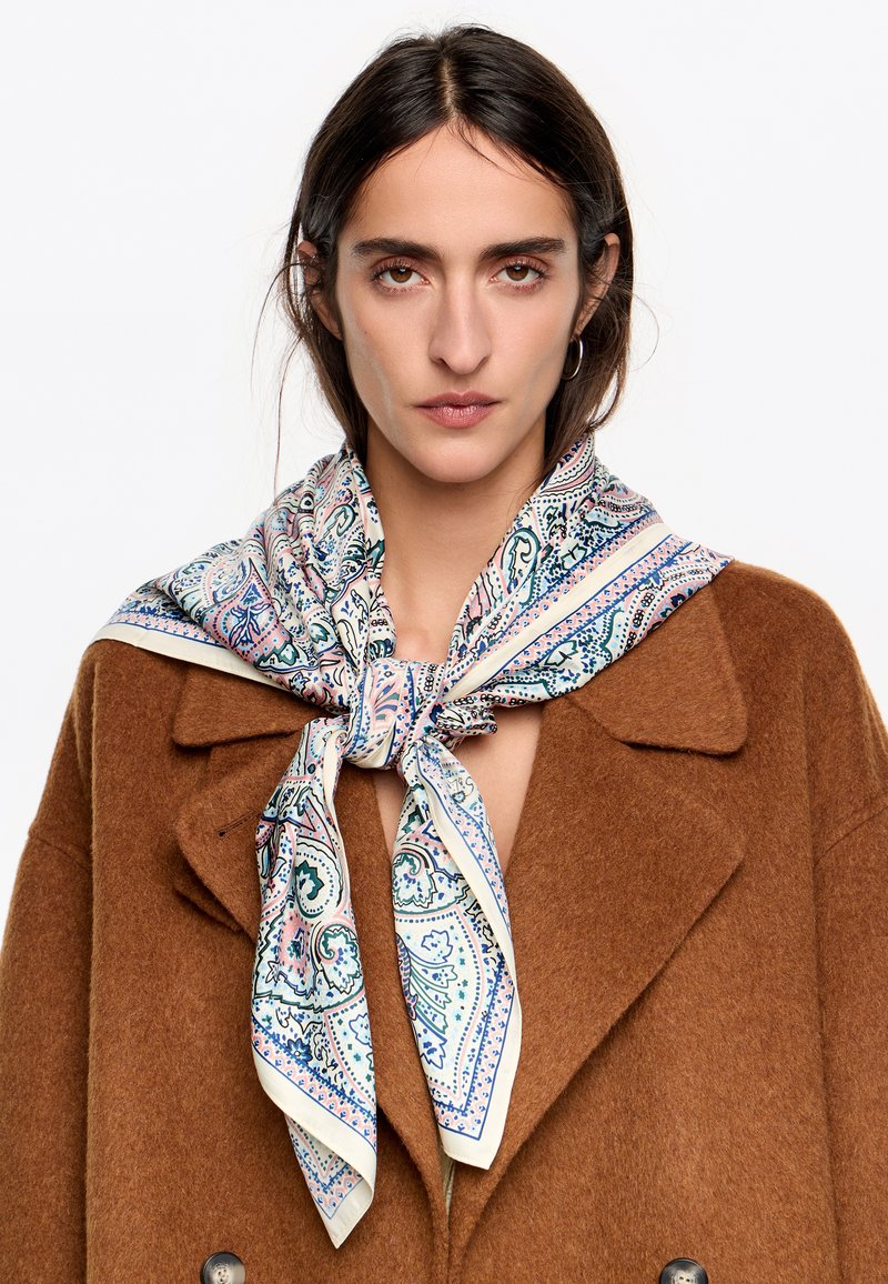 Jedwabny szalik w wzór paisley w kolorach niebieskim, różowym i kremowym, przewiązany wokół szyi na brązowym wełnianym płaszczu. Z wykończeniem w kremowej bordurze.