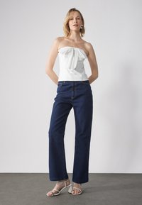 Hvit stroppeløs topp med sløyfedetalj, kombinert med mørkeblå vide jeans og hvite hæl-sandaler på et grått gulv.