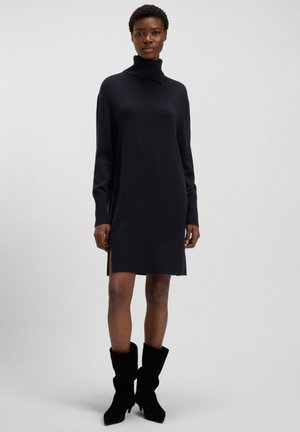 Frau in einem schwarzen Rollkragenpulloverkleid mit seitlichen Schlitzen und schwarzen Wildleder-Stiefeln mit Absatz bis zur Mitte der Wade, die vor einem schlichten Hintergrund steht.