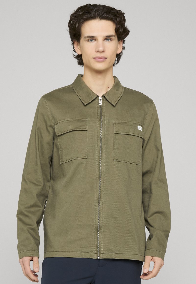 Veste zippée vert olive en tissu robuste, dotée de deux poches poitrine et d'un col classique. Texture lisse, détail de marque minimaliste.