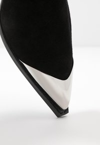 Zapato de tacón alto de ante negro con puntera afilada y un acento metálico plateado en la parte frontal. Superficie texturizada con un acento suave y brillante.