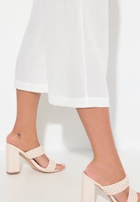 Sandales en beige tressées avec un talon épais, associées à un pantalon large texturé blanc s'arrêtant au milieu du mollet. Tatouage visible à la cheville.