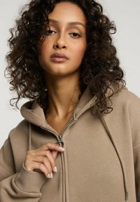 Beżowa bluza z kapturem na zamek, wykonana z miękkiego materiału, z kapturem ze sznurkiem i ściągaczami na mankietach. Tekstura wydaje się gładka, a krój jest luźny.