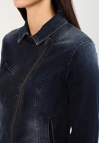 Dark blue denim jacket featuring a collared neckline, zip closure, side pockets, and contrasting stitching details.