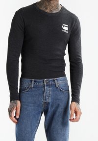 Pitkähiha, ympyräkaulainen toppi tummanharmaasta kankaasta, jossa on tiukka malli. Vasemmassa rinnassa on valkoinen logo. Yhdistetty sinisiin farkkuihin.