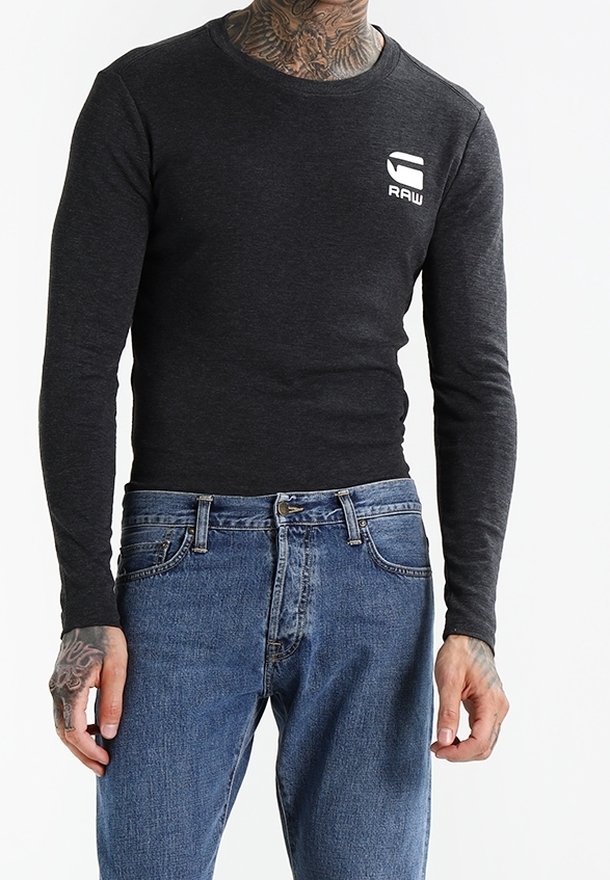 Pitkähiha, ympyräkaulainen toppi tummanharmaasta kankaasta, jossa on tiukka malli. Vasemmassa rinnassa on valkoinen logo. Yhdistetty sinisiin farkkuihin.