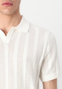 Camisa polo branca de malha com riscas verticais, apresentando um tecido texturizado, colarinho e mangas curtas, destacando um design respirável em trama aberta.