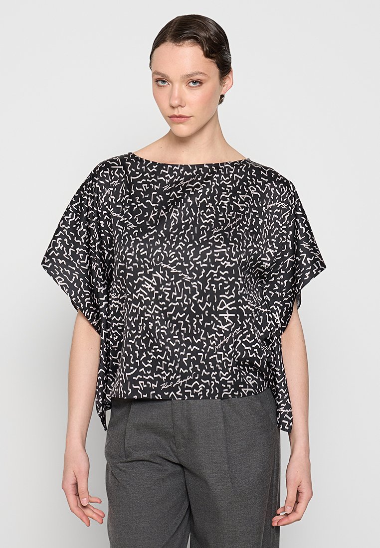 Haut noir avec des motifs abstraits blancs, manches courtes et larges, et une coupe décontractée. Associé à un pantalon gris, mettant en avant une silhouette décontractée.