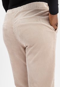 Beżowe spodnie kordurowe z pionowym prążkowanym wzorem, elastycznym pasem i dwoma tylnymi kieszeniami. Widok z boku podkreślający kształt i materiał.