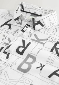 Balta krekla ar melnu grafisko rakstu, kas ietver burtus un formas, ar pogājamu apkakli un divām pogām priekšējā daļā.