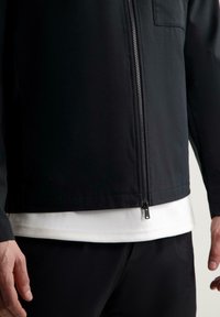 Giacca nera con zip realizzata in tessuto liscio, dotata di tasca frontale e di una camicia bianca a strati sotto. Design e struttura minimalisti.