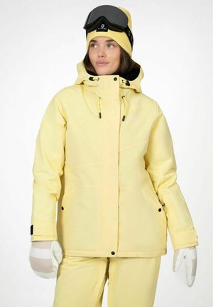 Kvinde iført en gul skijakke, matchende bukser, hvide handsker, en gul hue og sorte skibriller på panden.