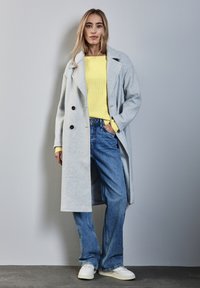 Ljusgrå dubbelknäppt kappa över en strukturerad gul tröja. Blå vida jeans och vita sneakers fulländar outfiten.