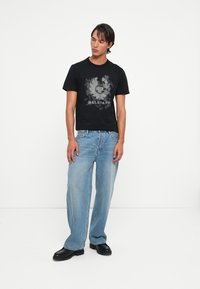 Črna majica s sivo grafično podobo na sredini, povezana z svetlo modrimi širokimi hlačami in črnimi debelimi čevlji. Enostavna bela ozadje.