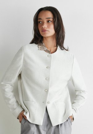 Kvinde med langt mørkt hår iført en tekstureret off-white knappet jakke over en mønstret skjorte, hænderne i lommerne, kigger til siden mod en ensfarvet baggrund.