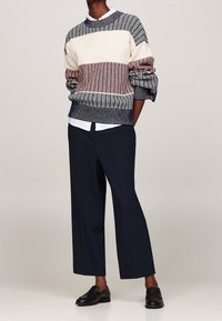 Gebreide trui met marineblauwe, crèmekleurige en bordeauxrode strepen, gedragen over een wit overhemd. Gecombineerd met marineblauwe wijde broekspijpen en zwarte loafers.