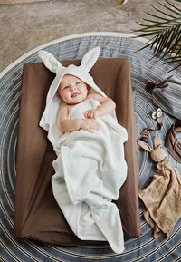 Valkoinen pupukorvatakki kiedottuna vauvan ympärille ruskealla patjalla, taustalla pyöreä, raidallinen peitto ja pehmeät lelut lähellä.