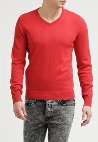 Pull-over rouge en v en tricot avec des manches longues, présentant des poignets et un ourlet côtelés, associé à un jean clair gris déchiré.