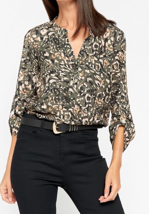 Vrouw die een blouse met camouflagepatroon draagt, met opgerolde mouwen, gestopt in een zwarte broek met een zwarte riem met een gouden gesp.