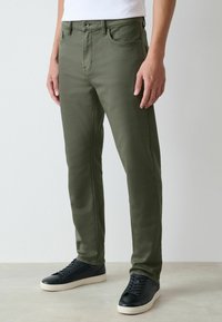 Oliivrohelised kitsas lõikega teksad, mis on valmistatud siledast puuvillakoorikust, viie taskuga, õmblustehnikaga ning nööbiga sulguriga. Kanda mustade spordijalanõudega.