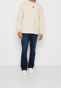 Persona che indossa un felpa beige con un piccolo logo, jeans blu scuro e sneakers bianche, in piedi davanti a uno sfondo bianco uniforme.