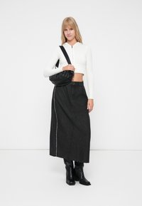 Pulover scurt alb cu fermoar, fustă maxi din denim negru cu cusături albe și cizme negre până la genunchi. Modelul poartă o geantă plic neagră.