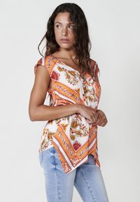 Blusa de manga corta con tela blanca y patrones florales y de paisley en naranja, bajo asimétrico y ajuste relajado. Combinada con jeans azules.