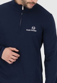 Pullover blu navy a mezza zip realizzato in tessuto liscio, con un logo bianco e il nome del marchio ricamato sul lato sinistro del petto. Maniche lunghe.