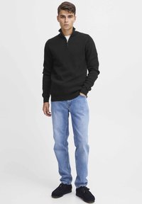Schwarzer Zip-Kragen-Pullover aus strukturiertem Strick, kombiniert mit hellblauen Jeans und schwarzen Mokkasins. Das Modell steht vor einem schlichten Hintergrund.