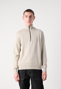 Sweater bege com fecho éclair e gola alta, punhos e bainha canelados. Apresenta um pequeno logótipo na poitrine. O material parece suave e liso.