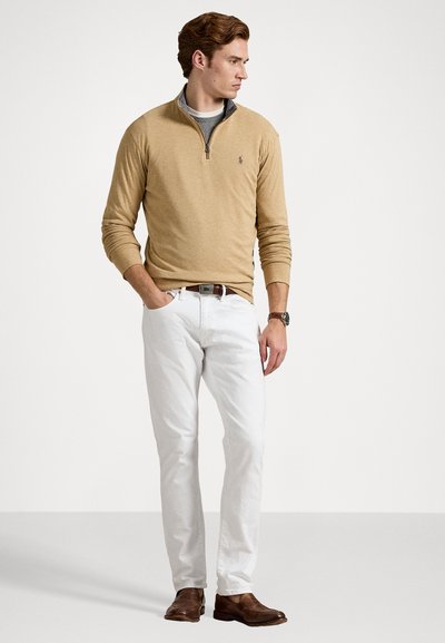 Bežinis puloveris su užtrauktuku, pilkas apykaklė, balti keliai ir rudi odiniai batai. Modelis stovi su rankomis kišenėse prieš baltą foną.