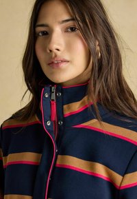 Hanorac bleumarin cu dungi orizontale îndrăznețe în bej și roz. Dispune de un guler înalt și un fermoar frontal cu un cursor metalic.