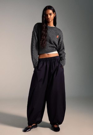 �Šedý pletený sveter s vzorom vrkoča a vyšívaným detailom, skombinovaný s čiernymi širokými nohavicami a čiernymi plochými topánkami.