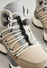 Beige a šedé turistické boty s texturovaným svrškem, elastickými šňůrkami a zámkovým systémem. Mají kulatou špičku a černou gumovou podešev.