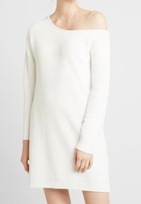 Biała dzianinowa sukienka o luźnym kroju, z jednym ramieniem, długimi rękawami, prążkowanym wykończeniem i teksturowanym materiałem.