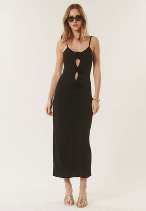 Femme portant une robe noire sans manches avec trois découpes devant, lunettes de soleil noires, bracelet en or et chaussures à talons ouverts dorés, debout devant un fond clair.