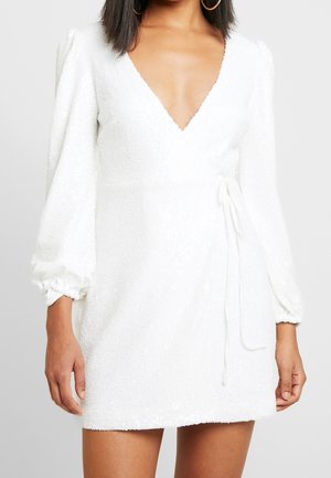 Frau trägt ein weißes, strukturiertes Mini-Wickelkleid mit langen Puffärmeln und tiefem V-Ausschnitt.