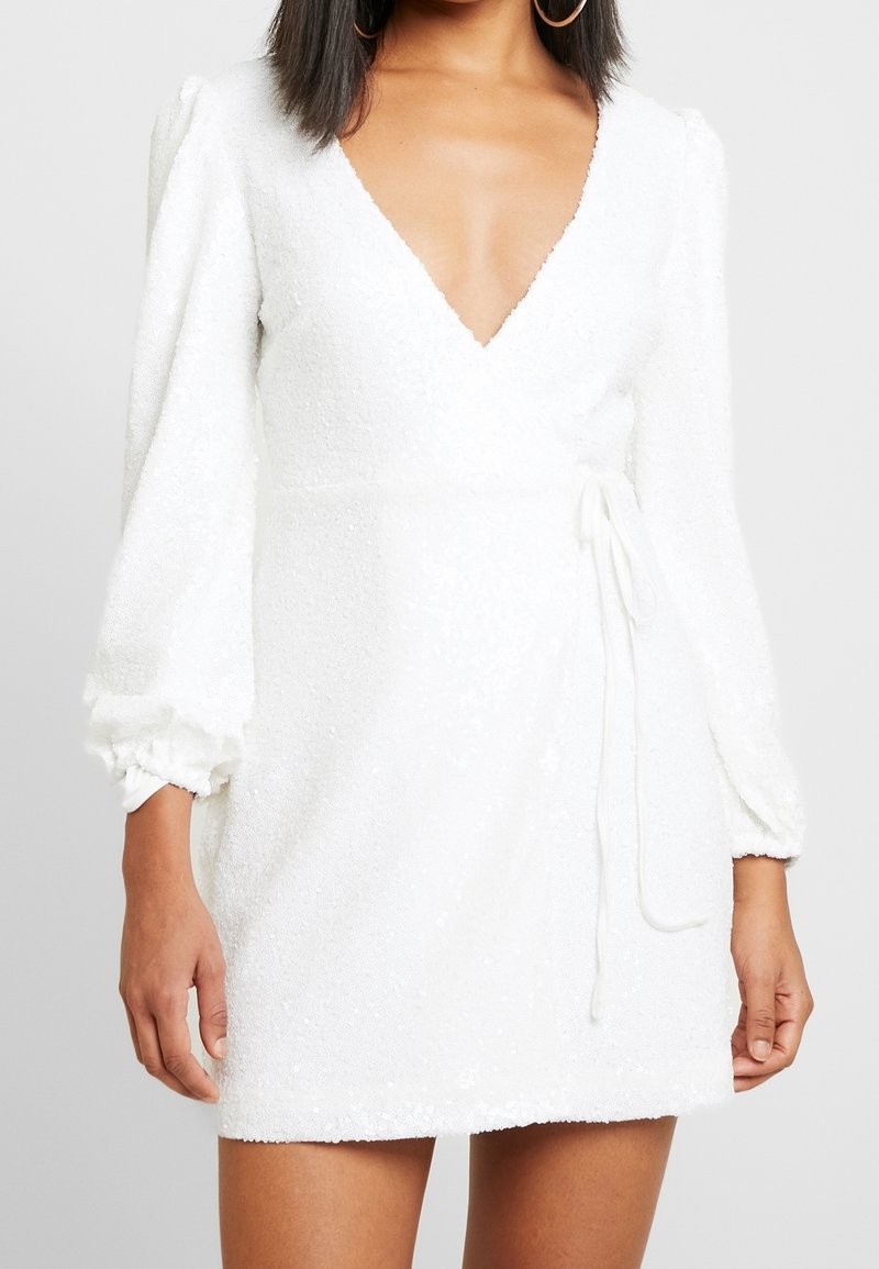 Femme portant une robe mini portefeuille blanche texturée avec de longues manches bouffantes et un décolleté en V profond.