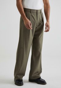 Pantaloni plisati verde măsliniu, cu o croială lejeră, având un fermoar și un sistem de închidere cu buton; purtați cu pantofi negri pe un fundal simplu.