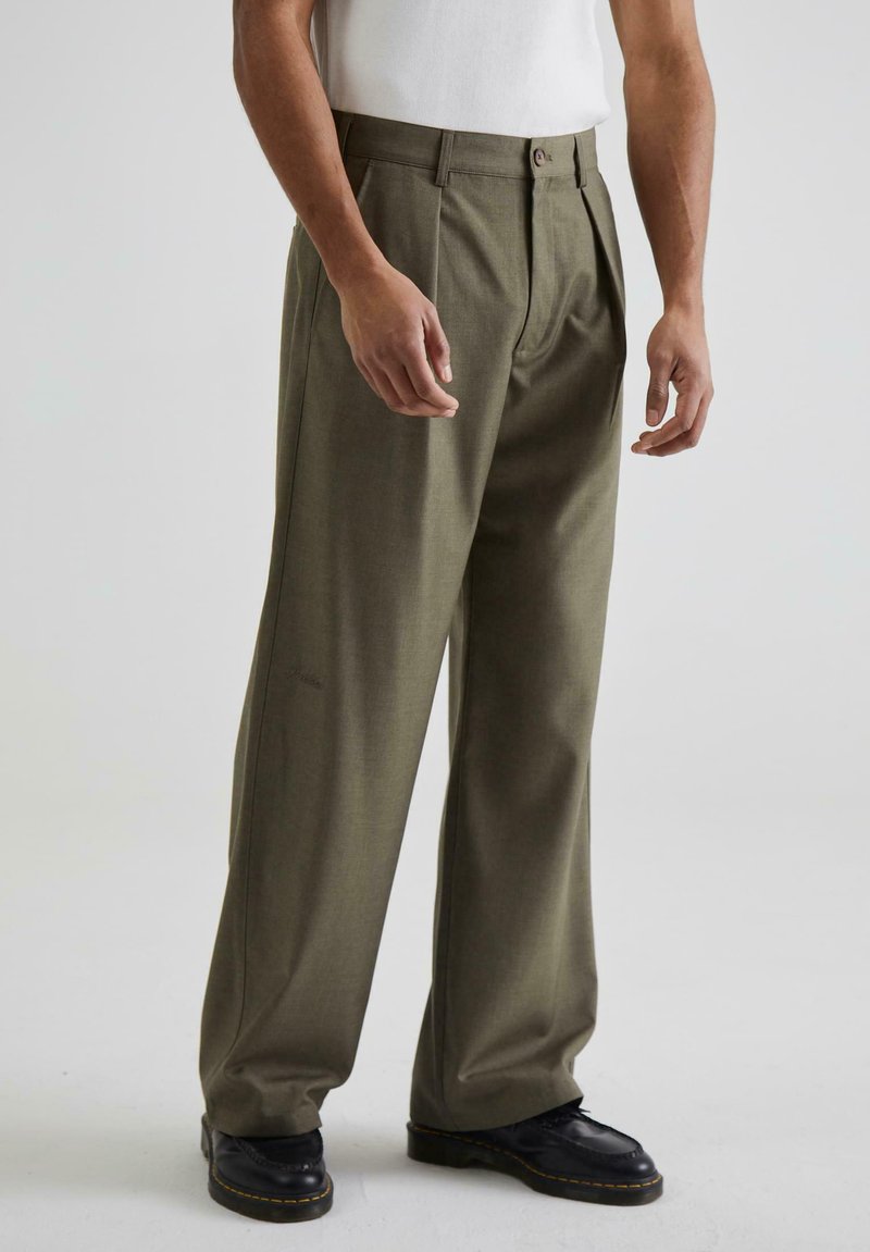 Pantaloni plisati verde măsliniu, cu o croială lejeră, având un fermoar și un sistem de închidere cu buton; purtați cu pantofi negri pe un fundal simplu.