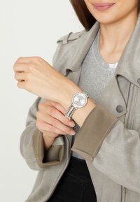 Montre argentée avec un cadran circulaire, un boîtier orné de pierres et un bracelet à maillons multiples. La tenue est composée d'une veste grise portée par-dessus un haut gris brillant.