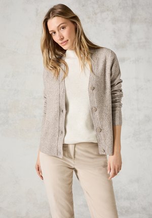 Gilet beige clair en laine douce, présentant une texture côtelée, des fermetures à boutons et des manches longues, porté sur un pull blanc et un pantalon beige.